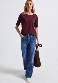 Bordeaux katoenen t-shirt met korte mouwen, gecombineerd met lichtblauwe spijkerbroek met wijde pijpen. Een bruine schoudertas en witte sneakers maken de look compleet.