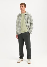 Man die een overhemd met patroon draagt over een lichtgroen T-shirt, donkere broek en witte sneakers, staand met één hand in zijn zak.