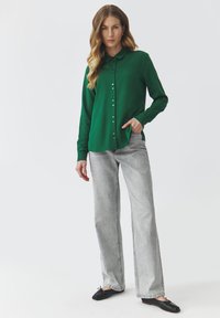 Green button-up shirt with a collar, long sleeves, and a relaxed fit; paired with light gray wide-leg jeans and black flats.