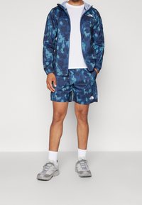 Herrträningsoutfit som består av en marinblå och teal camouflagemönstrad jacka och shorts, vit t-shirt, grå skor och vita strumpor. Lättviktsmaterial.