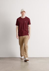 Jonge man die vooruitloopt en een beige pet, bordeauxkleurig t-shirt, kaki broek en witte sneakers draagt, tegen een effen witte muur.