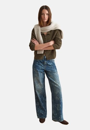 Vrouw met stijlbruin haar draagt een olijfgroen gebreid vest, een beige sjaal over de schouders gedrapeerd, wijd uitlopende blauwe spijkerbroek met bloemenprint en bruine schoenen.