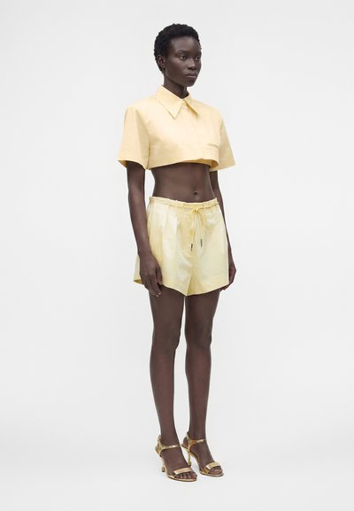 Femme portant une chemise courte à col jaune pâle, un short taille haute assorti et des sandales à talons hauts dorés, debout sur un fond blanc.