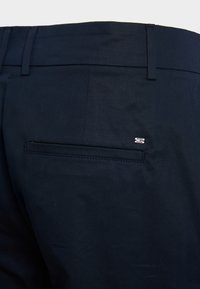 Mørkeblå bukser med en glat tekstur, som har en enkelt baklomme og et lille farvet logo ved kanten af lommen.