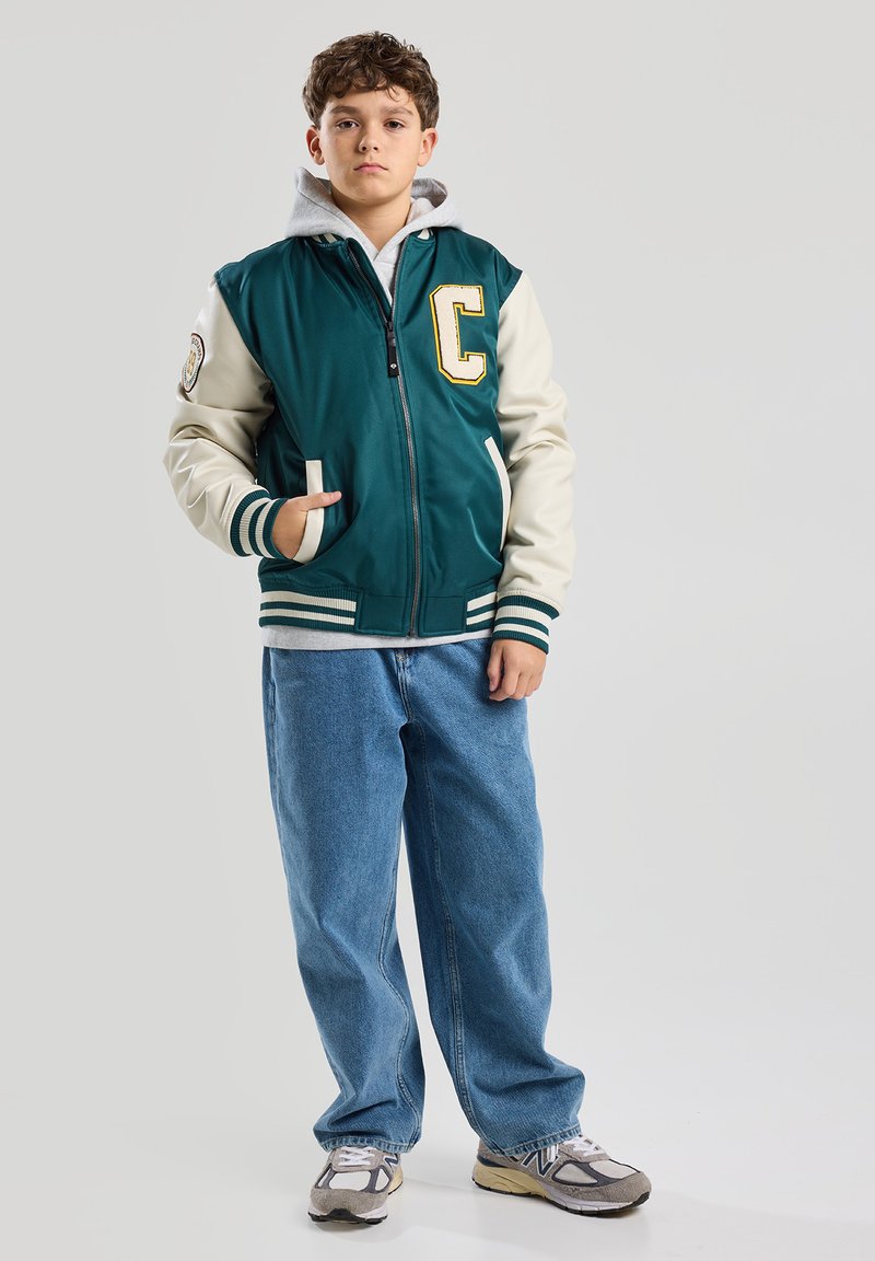 Green and cream varsity jacket with a letter “C” patch, white striped cuffs, and side pockets, worn over a gray hoodie and jeans.