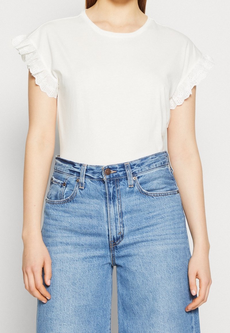Femme portant un haut blanc à manches courtes avec des manches à volants en dentelle et un jean bleu taille haute, mains détendues le long du corps.