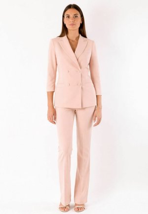 Donna che indossa un blazer doppiopetto color rosa chiaro con maniche a tre quarti e pantaloni a gamba dritta abbinati, in piedi con espressione neutra su sfondo bianco.