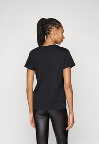 T-shirt nera in cotone a maniche corte, con scollo rotondo e vestibilità rilassata, vista da dietro, abbinata a leggings neri lucidi.