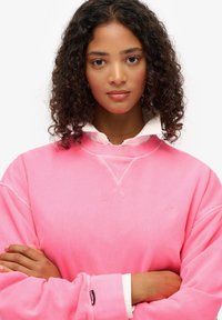 Sudadera rosa con corte relajado y mangas ligeramente exageradas, con un pequeño logo y superpuesta sobre una camisa blanca con cuello.