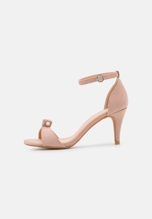 Beige ruskind sandal med høj hæl, ankelrem, åben tå og lille perledetalje på frontremmen.