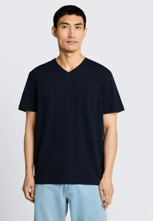 Man met kort zwart haar die een effen donkerblauw V-hals T-shirt en lichtblauwe spijkerbroek draagt, staand tegen een effen achtergrond.