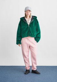 Πράσινη μαλακή ζακέτα με φερμουάρ και κουκούλα, λευκό πουκάμισο με κουμπιά, ροζ φόρμες και μαύρα αθλητικά παπούτσια. Μπεζ καπέλο με λογότυπο. Απλό φόντο.