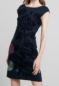 Czarna sukienka o dopasowanym kroju, z misternym haftem floralnym w żywych kolorach. Rękawy typu cap oraz okrągły dekolt dopełniają całość.