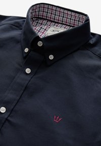 Camicia blu navy scura con bottoni e colletto ripiegato, con logo ricamato rosa. Motivo a quadri visibile sulla fodera interna del colletto. Realizzata in tessuto liscio.