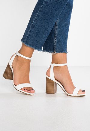 Sandales à talons blancs avec deux larges brides et une fine bride au niveau de la cheville. Talon bloc marron structuré avec un revêtement vertical et un design à bout ouvert.