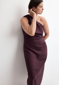 Femme en robe asymétrique sans manches de couleur violet foncé, debout de profil, touchant ses cheveux, devant un mur blanc uni.