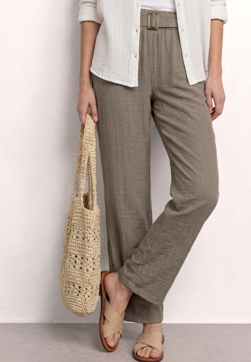 Mujer que lleva pantalones beige de pierna ancha con cinturón, camisa blanca, sandalias marrones, sujetando un bolso Tote beige de crochet.