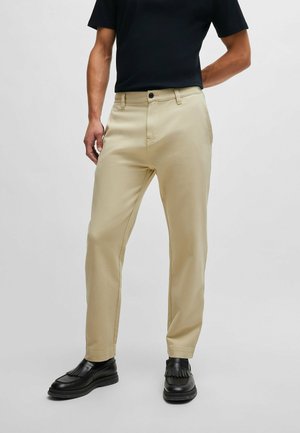 Beige chinos with a straight-leg fit, featuring a zip fly, button closure, and front pockets. Paired with black loafers.