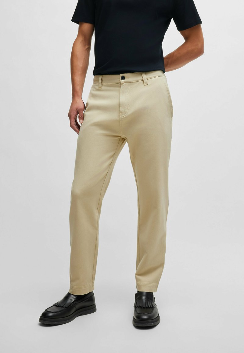 Beige chinos with a straight-leg fit, featuring a zip fly, button closure, and front pockets. Paired with black loafers.