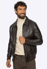 Bruine leren bomberjack met een kraag, geribbelde manchetten en een rits aan de voorkant. Gedragen over een lichtbeige coltrui en bruine broek.