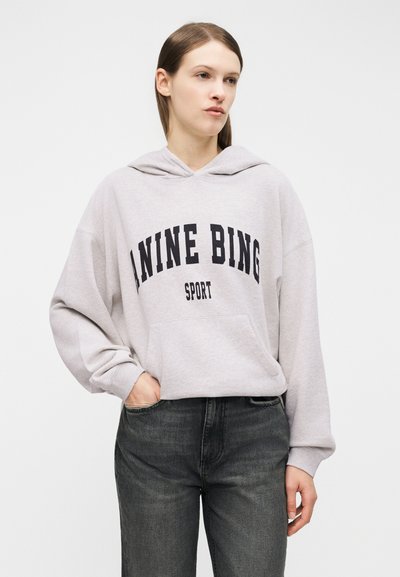 Young woman wearing light grey hoodie with "ANINE BING SPORT" text and dark gray jeans, standing with one hand in pocket.
