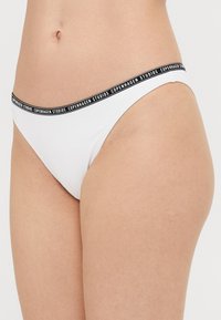 Hvite bikiniunderdeler med lav midje-fit, med et svart elastisk belte med "COPENHAGEN STUDIOS" trykket langs kanten.