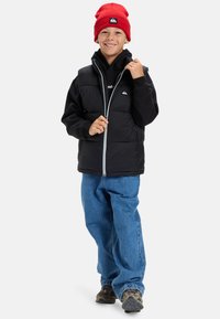 Leende pojke bär en röd mössa, svart puffer-väst över en svart huvtröja, lösa blå jeans och bruna sneakers, stående mot en vit bakgrund.