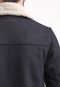 Svart ullkappa med en krämfärgad fårskinnskrage. Har rena linjer, synliga sömmar och en liten röd broderad detalj på baksidan.