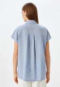 Camisa a rayas azules con mangas cortas tipo cap sleeve, un pliegue en la parte trasera y un cuello clásico. La tela parece ligera y tiene una textura suave.