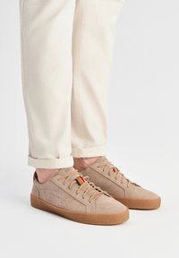Baskets en daim beige clair avec une finition texturée, semelle en caoutchouc et accent décoratif à rayures sur la languette ; associées à un pantalon beige clair retroussé.