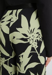 Black fabric with large, abstract light green floral and leaf patterns, featuring a smooth texture and a black satin waistband.