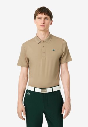 Giovane uomo che indossa una polo beige e pantaloni verde scuro con una cintura bianca, in piedi davanti a uno sfondo bianco uniforme.