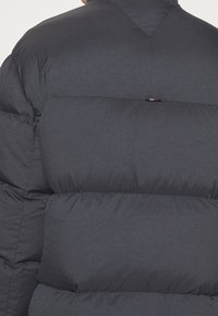 Ciemnoszara kurtka puchowa z pikowanymi sekcjami, wyposażona w wysoki kołnierz oraz małe logo w kolorach czerwonym, białym i niebieskim na plecach.