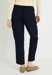 Pantalones azul marino con cintura elástica, diseño de corte ajustado y bolsillos traseros; combinados con zapatos planos beige con tachuelas decorativas.