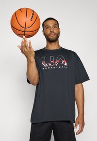 Sort kortærmet T-shirt med rød og hvid "UA BASKETBALL" grafikdesign, båret af en person der holder en basketball, mod en ensfarvet baggrund.