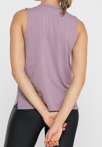 Lavendelfärgad ärmlös topp med avslappnad passform, tillverkad av mjukt tyg; har rundad halsringning och sidofickor. Bärs med svarta, tighta leggings.