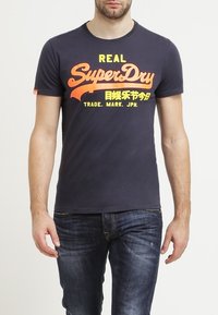 Námořnicko modré bavlněné tričko s velkým žlutým a oranžovým textem "REAL SuperDry" a grafickým logem. Kulatý výstřih, krátké rukávy, volný střih.