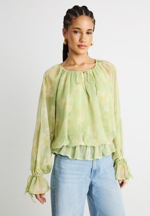 Nainen, jolla on punottu tukka, kullatut korvakorut ja vaaleanvihreä kukkakuosinen pitkähihainen paita sekä vaaleansiniset farkut valkoista taustaa vasten.