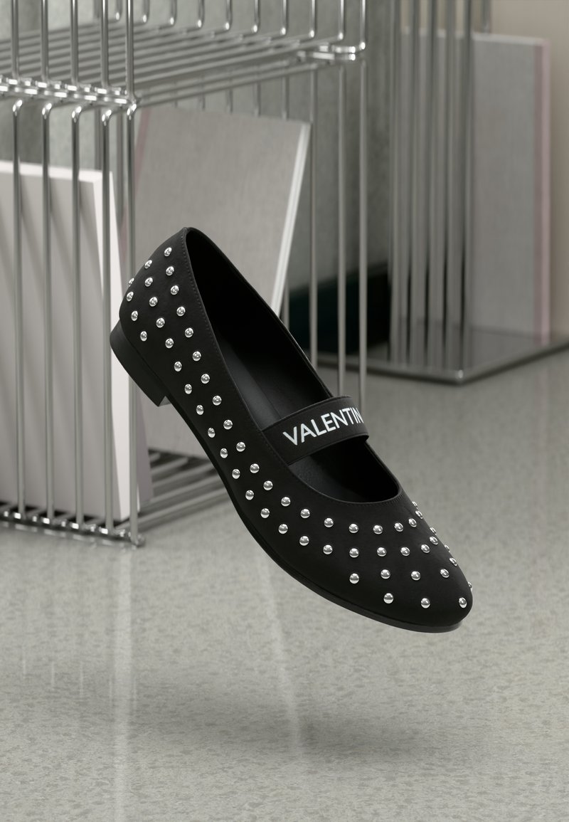 Zapato plano negro con acentos de tachuelas plateadas, acabado suave y una correa con la marca que lleva el nombre "VALENTINO". Diseño elegante y de perfil bajo.