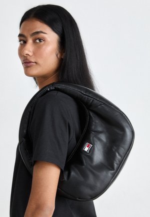 Kobieta o długich czarnych włosach, ubrana w czarną koszulę, nosząca na ramieniu zakrzywioną czarną skórzaną torbę z małym prostokątnym logo.