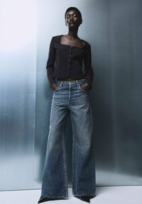 Top negro ajustado de manga larga con escote cuadrado y botones, combinado con jeans de mezclilla azul de pierna ancha que presentan detalles brillantes.