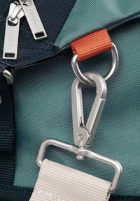 Un mousqueton en métal attaché à un tissu texturé vert et bleu marine avec des détails de sangle orange. Deux fermetures éclair argentées sont visibles en haut.