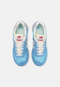 Blå sneakers i mocka och mesh-material, med vita detaljer, en grå tunga och en röd etikett. Modellnummer 574 på sidan.