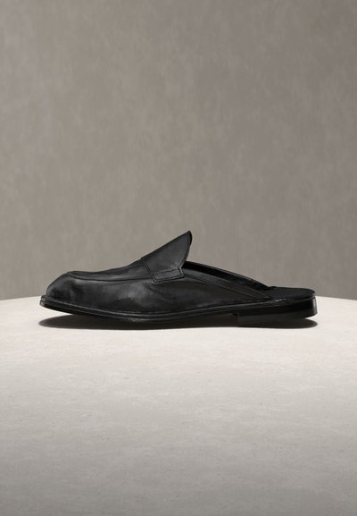 Μαύρο δερμάτινο μοκασίνι χωρίς κορδόνια, με ανοιχτή φτέρνα, στρογγυλή μύτη και χαμηλό τακούνι, εμφανίζεται σε ανοιχτή επιφάνεια με ουδέτερο φόντο.