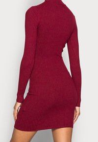 Abito a maglia a coste in bordeaux, con colletto alto, maniche lunghe e silhouette aderente. Tessuto liscio con lunghezza fino a metà coscia.