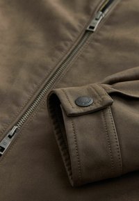 Brown suede jacket sleeve with buttoned cuff resting near a metal zipper on the front of the jacket.
