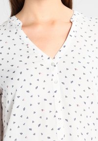 Blusa bianca con scollo a V e bottoni, caratterizzata da un delicato motivo di piccole foglie blu e rosa. Tessuto leggero e traspirante.