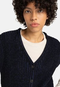 Donkerblauwe cardigan met een ribbelstructuur, voorzien van een V-hals, drie knopen en een lichtgewicht stof, gedragen over een crèmekleurig shirt.