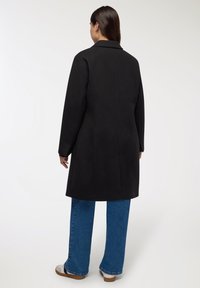 Donna in piedi di spalle, che indossa un cappotto nero fino al ginocchio, jeans blu e sneakers bianche su uno sfondo chiaro e uniforme.