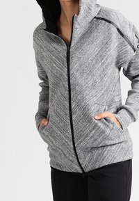 Sudadera gris con textura, con cremallera y detalles en negro, capucha y bolsillos laterales. Presenta un diseño aerodinámico y material suave y elástico.